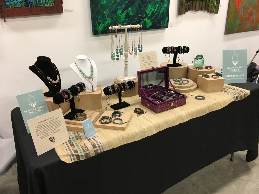 Display table with handmade jewelry at a crafts show, with paintings on the wall in the background.